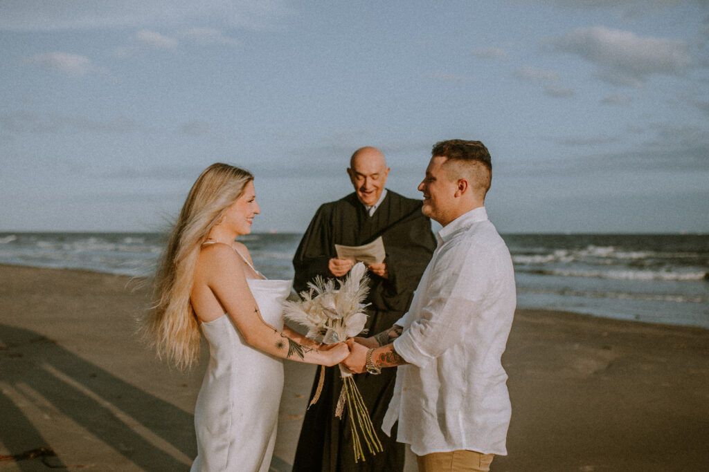 Couple holding hands, at the beach, while a judge gets them married.