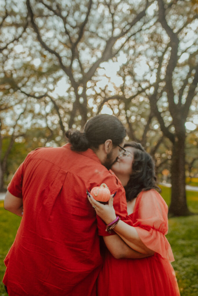 Couple kissing and holding a small pumpkin during their maternity photoshoot.