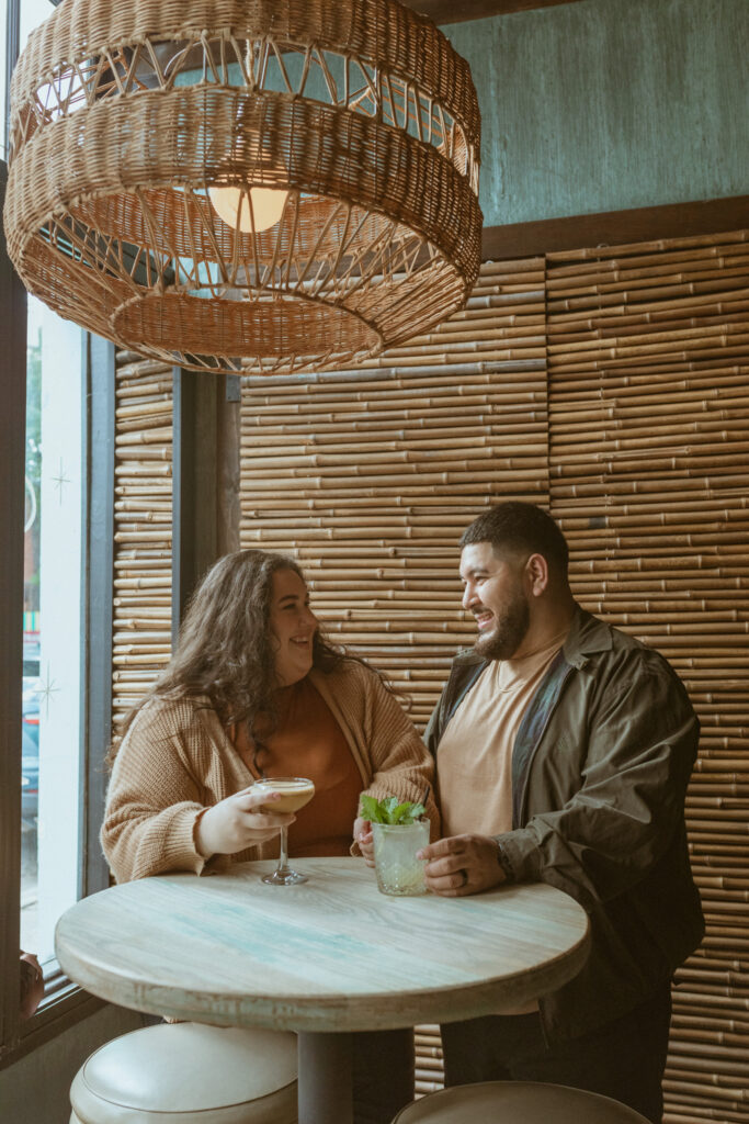 Couple laughing sitting at a table, having drinks, during their 1st Year Anniversary Photoshoot 