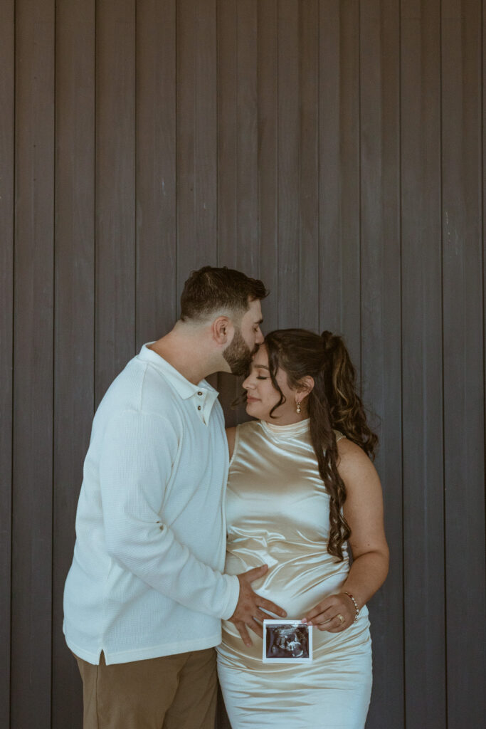 Man kissing his wife on the forehead while woman hold their baby's ultrasound during their maternity photoshoot.