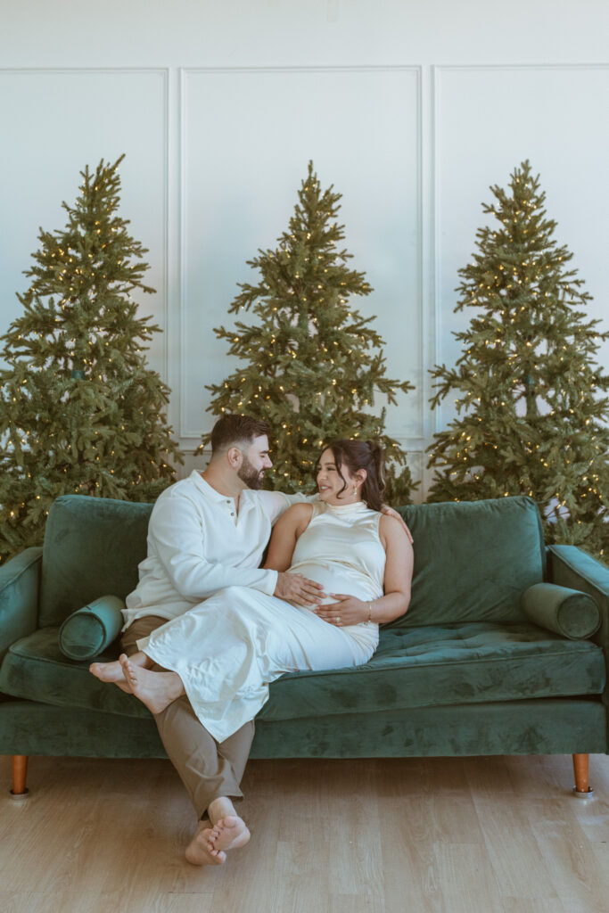 Couple sitting on a couch with christmas trees in the back during their maternity photoshoot.