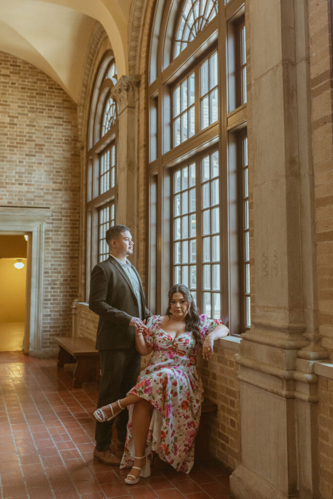 man looking out the window while woman, wearing a floral dress, during their engagement photos at Julia Ideson Library