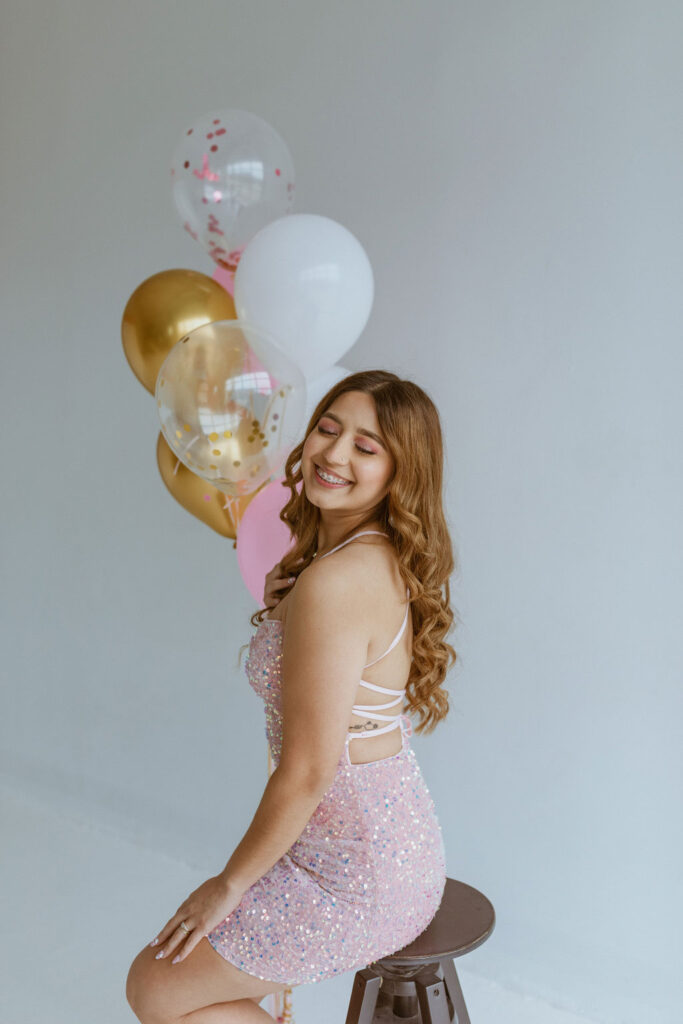 Girl wearing a pink dress sitting on a stool with balloons ins the background.