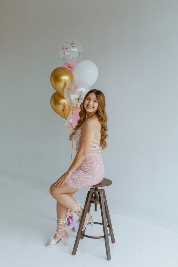 Girl wearing a pink dress sitting on a stool with balloons ins the background.