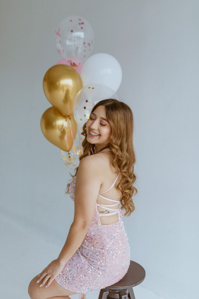 Girl wearing a pink dress sitting on a stool with balloons ins the background.