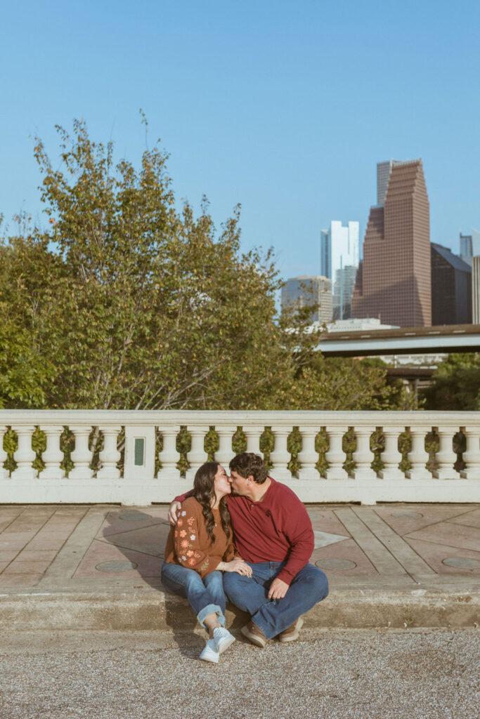 Couple sitting on a sidewalk, with the Houston Skyline in the background, during their fall photoshoot. 